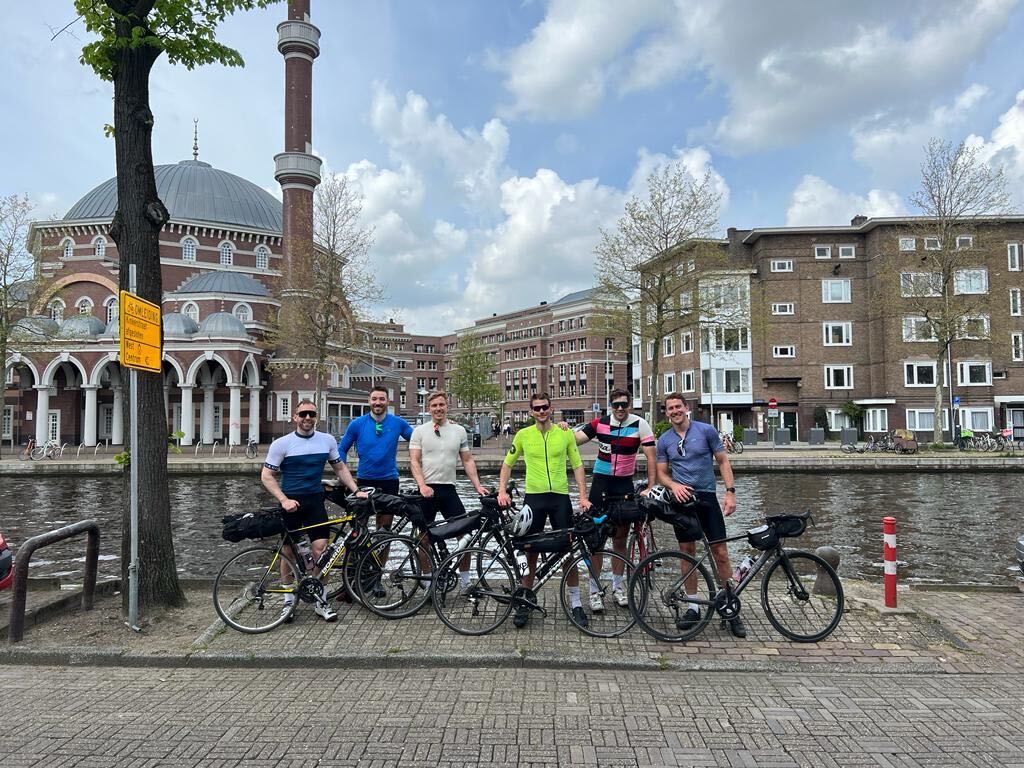 Group of cyclists with bikepacking setups beside an Amsterdam canal at the start of a multi-day European cycling tour